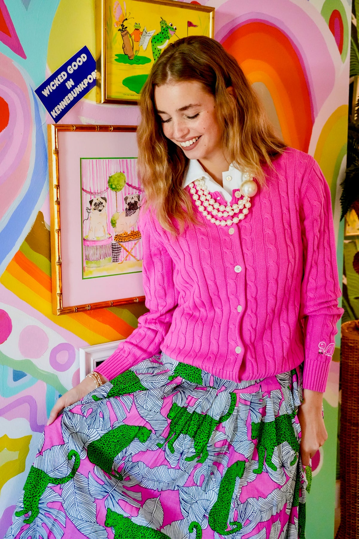 Woman in a pink cardigan and colorful skirt standing in front of a vibrant, abstract wall with framed pictures.