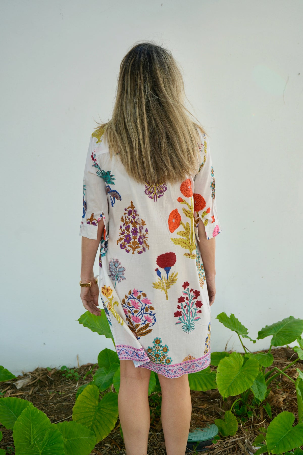 Person wearing a floral dress standing in a garden with a white wall in the background