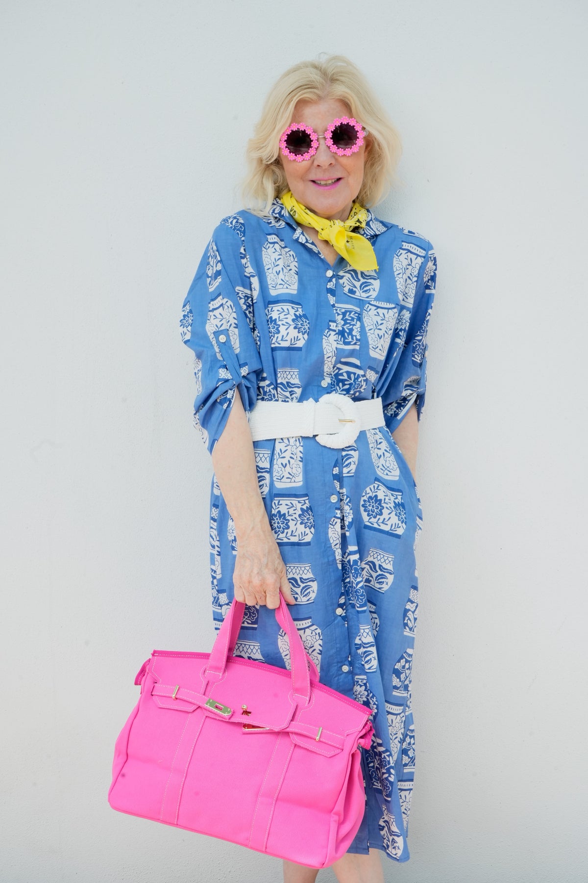 Woman in a blue dress with white patterns, holding a pink bag, wearing pink sunglasses, and a yellow scarf against a plain background.