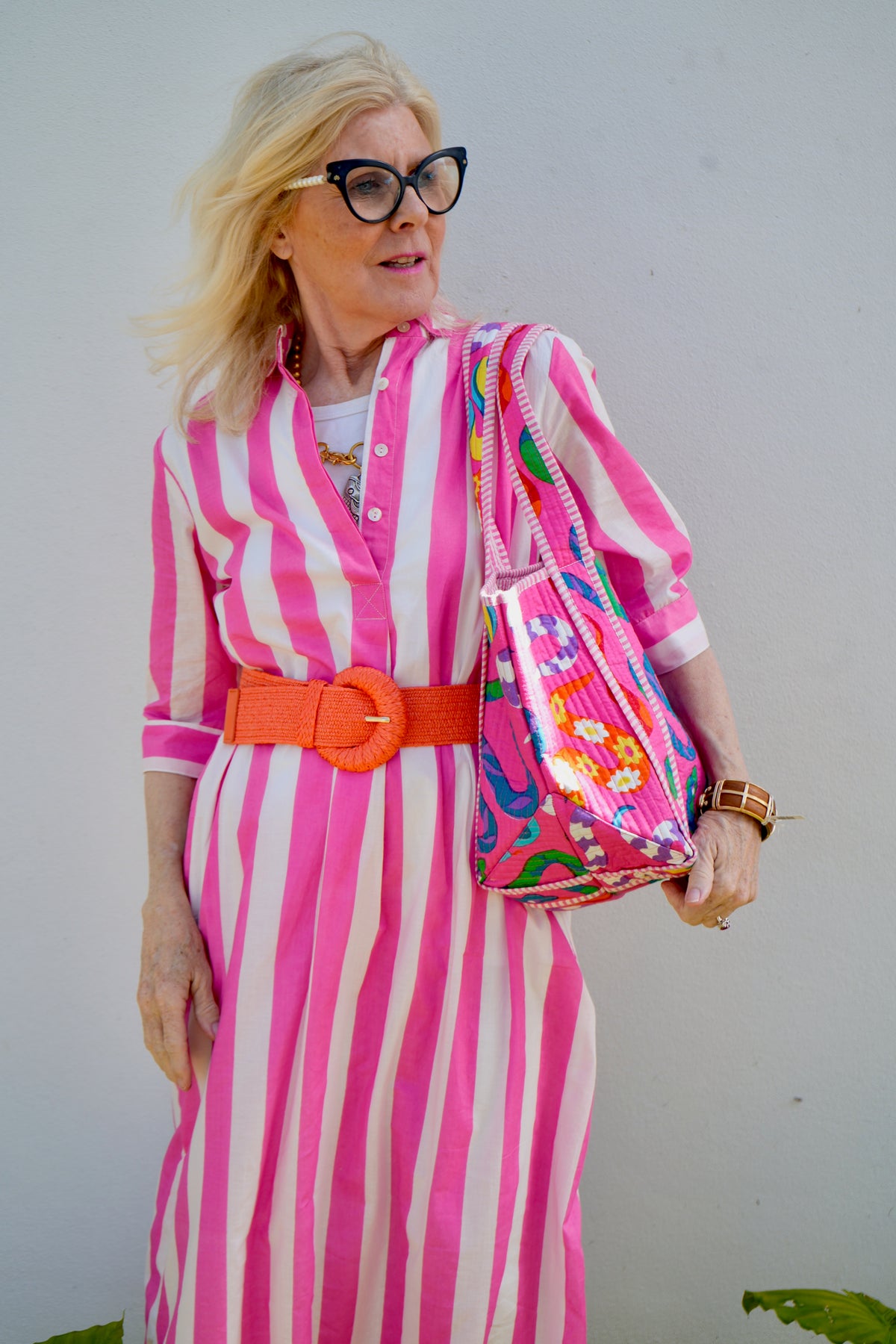 Woman in a pink and white striped dress with an orange belt, holding a colorful bag against a plain background.