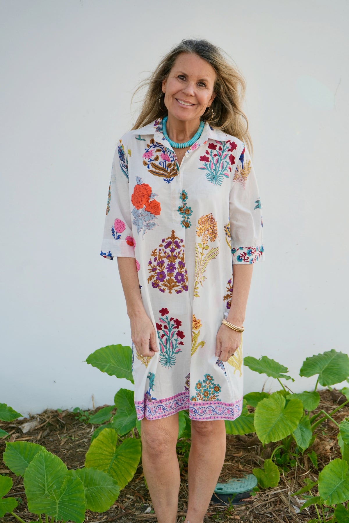 Woman wearing a colorful floral dress standing outdoors with greenery around.
