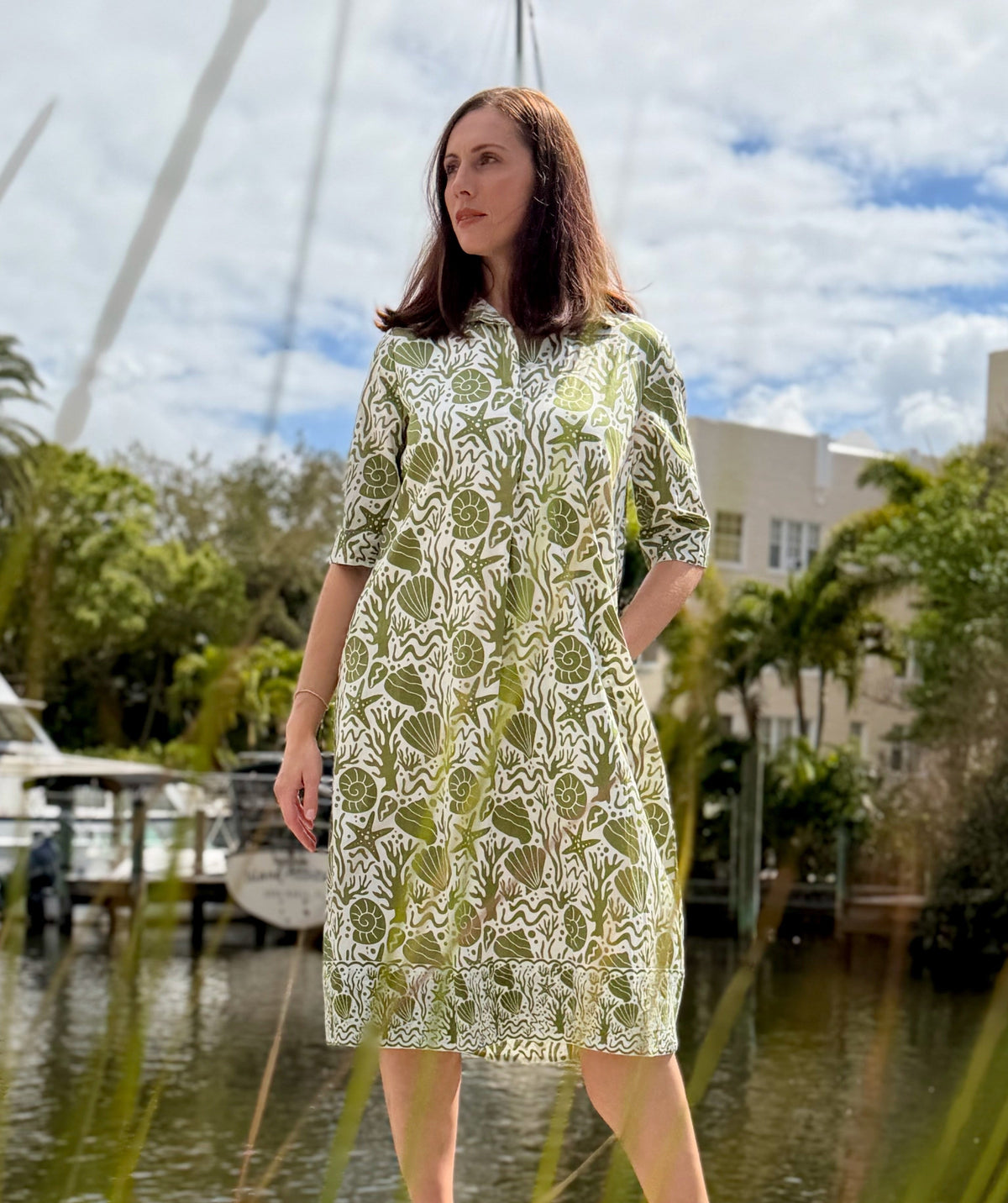 Woman in a green and white dress standing by a waterfront with boats and trees in the background.